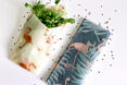 Two folded beeswax wraps with flamingo patterns, one with cilantro inside and one without on a white surface sprinkled with brown seeds