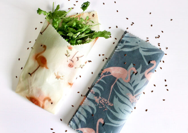 Two folded beeswax wraps with flamingo patterns, one with cilantro inside and one without on a white surface sprinkled with brown seeds