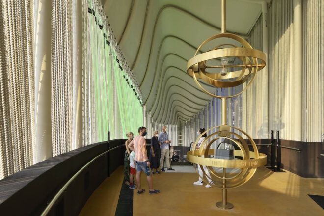 A rendering of people walking through a pavilion and looking at a structure with two golden globe-like fixtures.