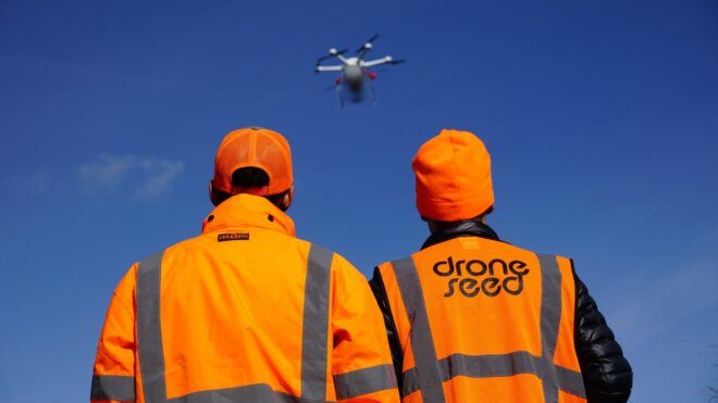 Two men wearing orange caution jackets watching a drone fly