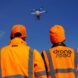 Two men wearing orange caution jackets watching a drone fly