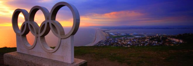 A gray statue of the Olympic rings in front of a coastal landscape.