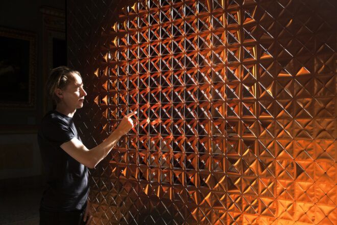 A person pointing a finger at a gold wall that is designed with multiple holes