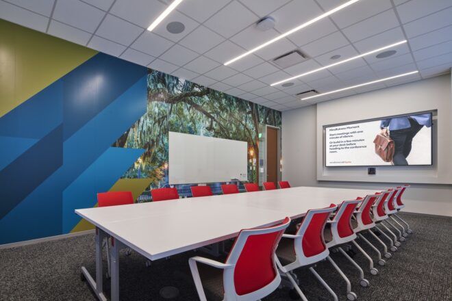 A room with a long rectangle table with chairs on both sides of it and a monitor on the wall