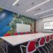 A room with a long rectangle table with chairs on both sides of it and a monitor on the wall