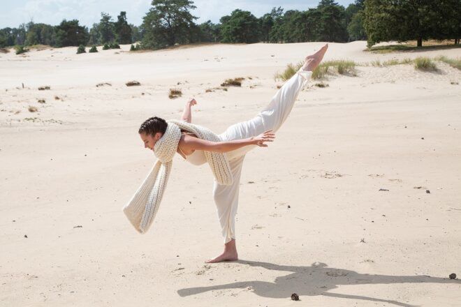 A woman balancing on one leg wearing a white outfit with a white wool scarf