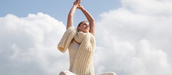 A woman sitting cross-legged with her hands clasped above her head sitting in sand area wearing a white wool scarf and white outfit