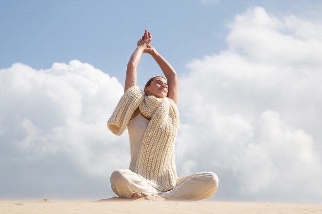 A woman sitting cross-legged with her hands clasped above her head sitting in sand area wearing a white wool scarf and white outfit