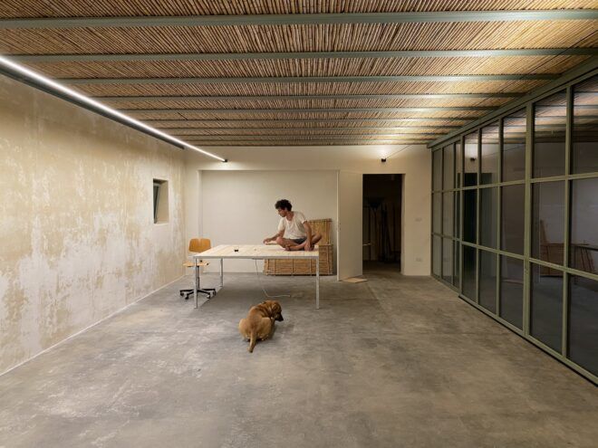 A room with a desk and a person sitting on the desk with a dog on the ground and floor-to-ceiling windows on the right