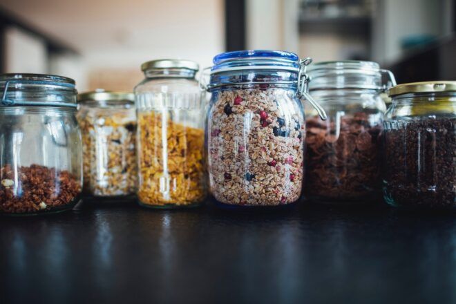 Jars on a black counter filled with dried pasta, nuts and other assortments of food