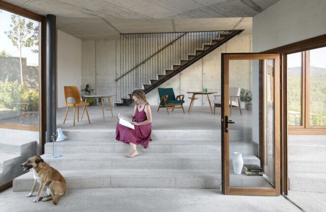 A woman sitting on stone steps inside and a dog looking out the window. There is also a staircase behind her