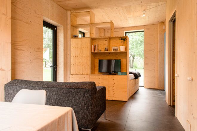 View across a desk towards a living space with shelving and a bed at the back facing a floor-to-ceiling window