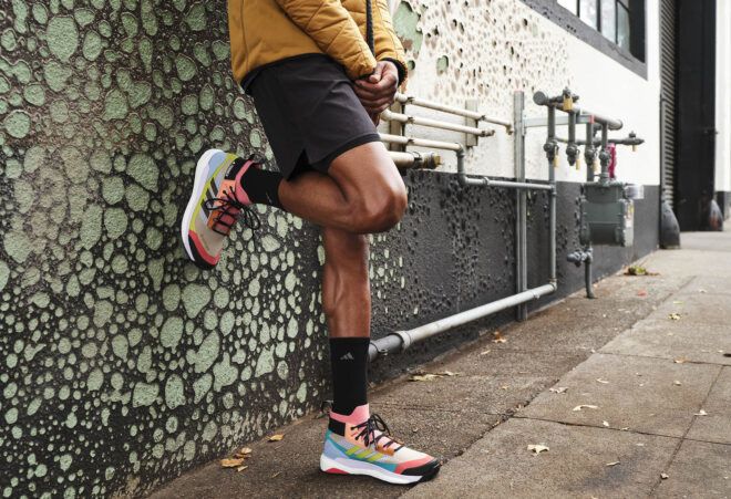A pair of legs against a wall, wearing mulitcolor Adidas shoes.