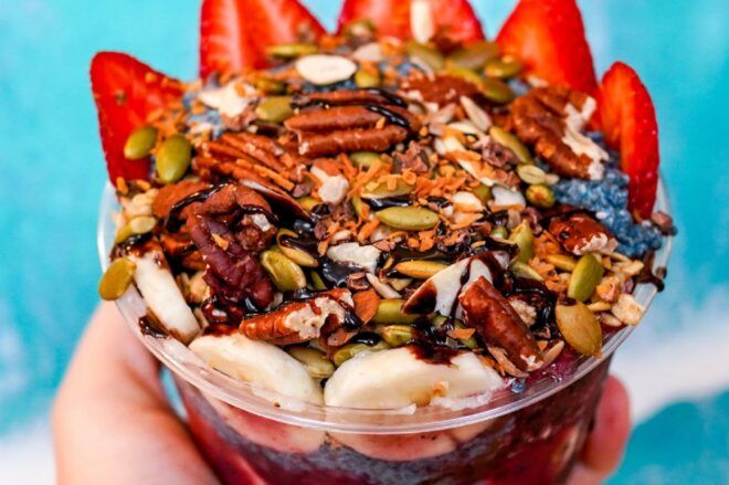 A hand holding a smoothie bowl loaded with nuts, strawberries and banana slices against a blue background