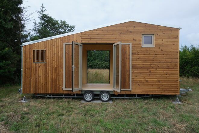 A tiny wooden house on wheels sits in a grass field