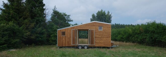 A tiny wooden house on wheels sits in a grass field