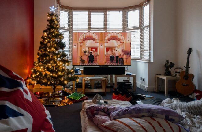 A Christmas tree in the corner of a living room beside a TV turned on