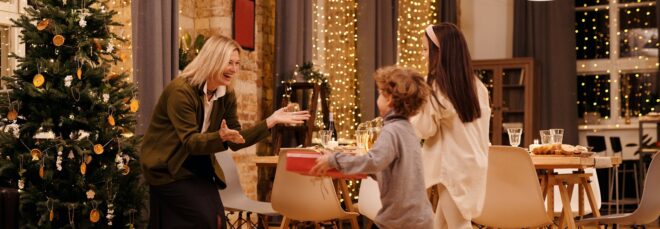 A woman standing in front of a Christmas tree with her arms outstretched to two children who are running toward her with presents