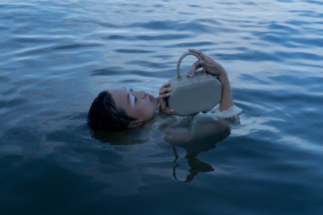 A woman submerged underwater except for her head and arms holding a blue handbag above the water
