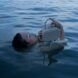 A woman submerged underwater except for her head and arms holding a blue handbag above the water