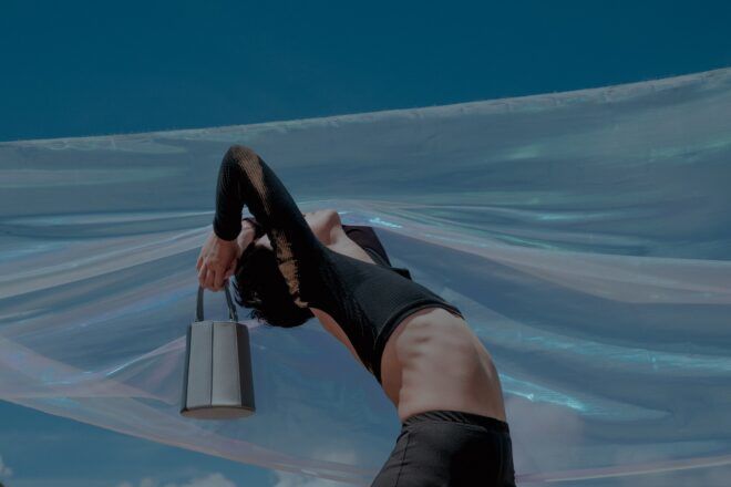 A woman bending backwards holding a handbag bucket behind her with a sheer cloth flowing above her body