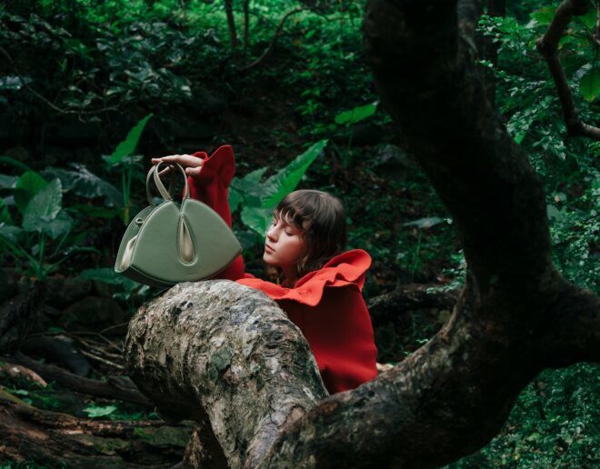 A woman wearing an orange top leans against a tree bark with a green handbag placed on the branch of the tree