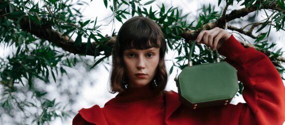 A woman with an orange top and hair cut above her shoulders holds a green handbag to rest on her shoulder
