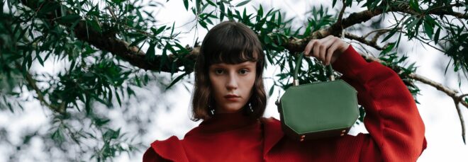 A woman with an orange top and hair cut above her shoulders holds a green handbag to rest on her shoulder