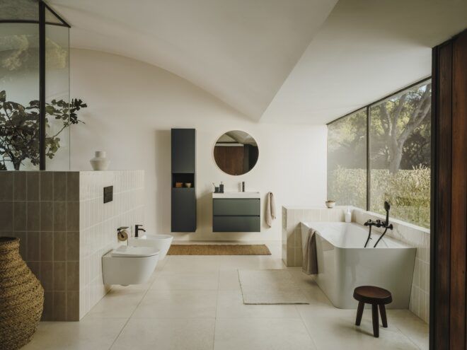 A minimalistic bathroom that has a white bathtub on the right that is against a window, a sink area with a circle mirror above it and another tiled area to the left