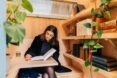 A woman sits on a bench with a fold out table where she is writing in a journal and next to her are bookshelves