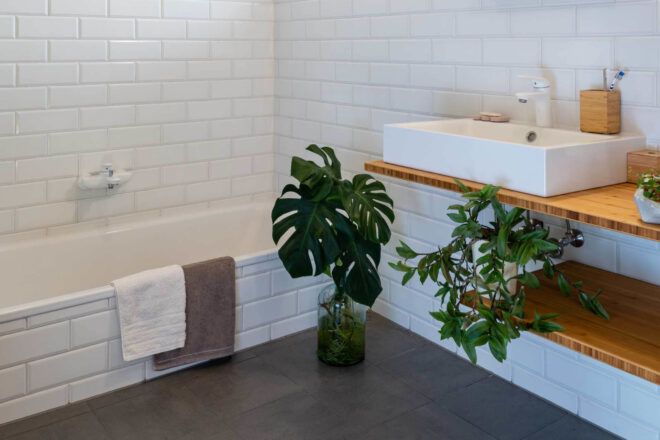 White tile-clad bathroom with a tub, sink, and plants on shelves and on the grey tiled floor