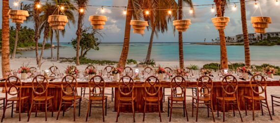 A large rectangle table set in the beach with chairs filling it on either side and wooden chandeliers hanging above it