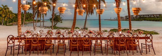 A large rectangle table set in the beach with chairs filling it on either side and wooden chandeliers hanging above it