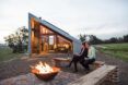 Two people sitting in front of a bonfire in front of a tiny house with a sloped roof.