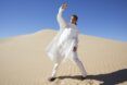 A woman with her hand up to shield from the sun is standing in a desert wearing a white outfit