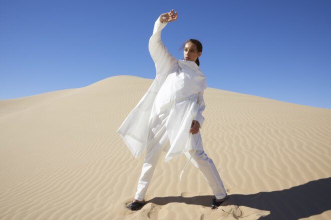 A woman with her hand up to shield from the sun is standing in a desert wearing a white outfit
