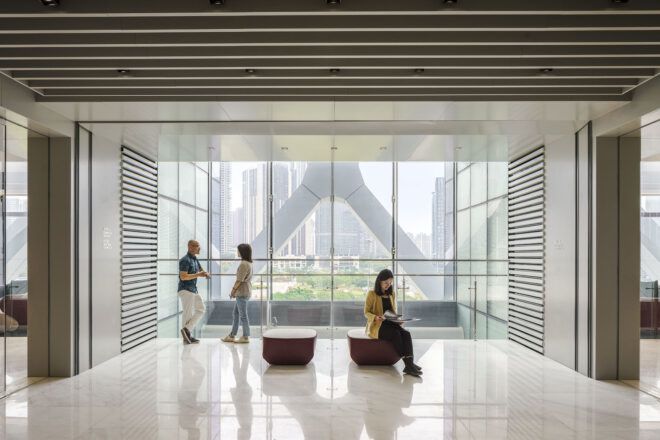 A lounge area where people sit and talk by a wall of windows.