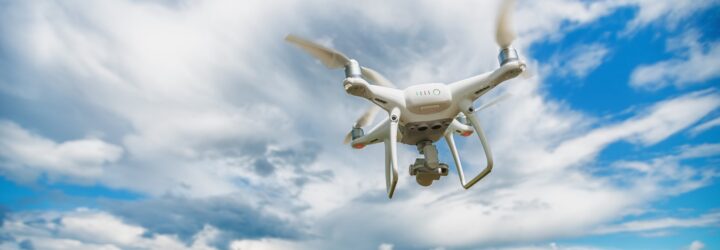 A flying white drone across a blue sky with white clouds