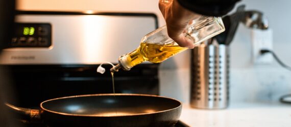 A hand pouring oil in a glass container into a black pan on a stove