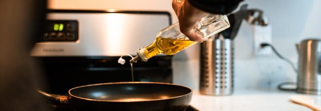 A hand pouring oil in a glass container into a black pan on a stove