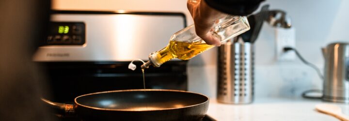 A hand pouring oil in a glass container into a black pan on a stove