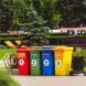 Four bins in red, green, blue and yellow colors. They sit on the pavement in a park