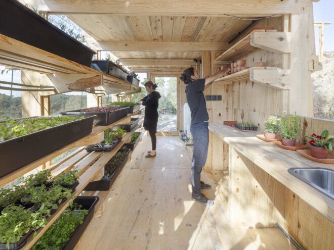View of the interior of the greenhouse's ground level. A woman faces the shelving units on the left while a man stacks items on the shelf above the counter on the right