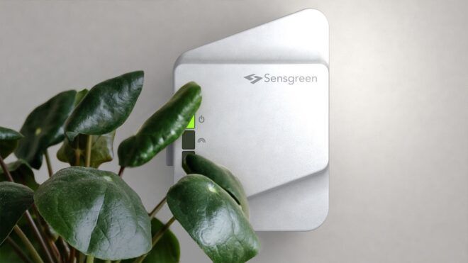 A white Sensgreen mounted on a wall with a plant in front of it