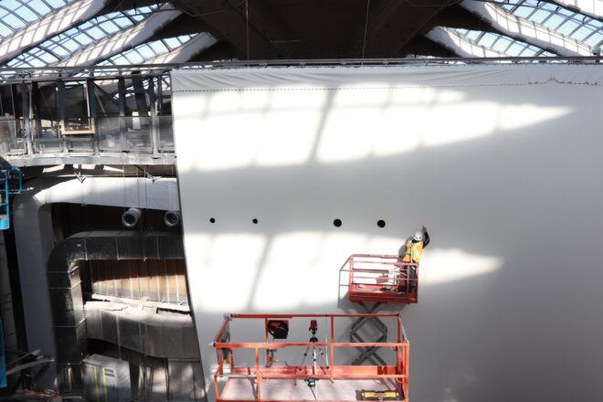 A large white wall where a man standing on an orange structure to fix something on the white wall