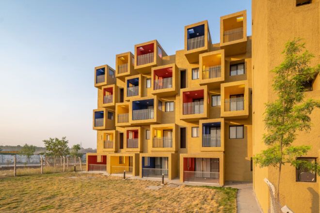 A yellow ochre sustainable housing building with colorful cube-shaped apartments that cantilever off the structure, with a flat garden space at the front