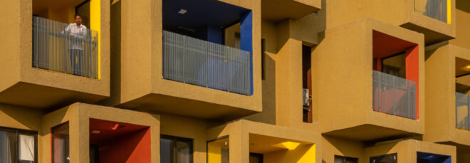 A close up of a yellow ochre building with colorful cube-shaped apartments that cantilever off the structure. Two male residents can be seen peering off their balconies