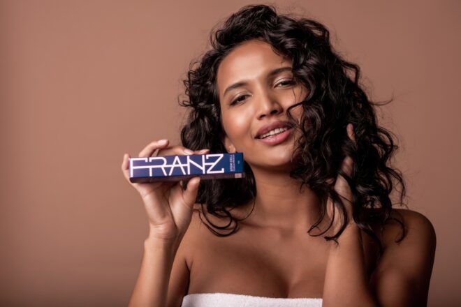 A brown-skinned woman holding a slim packaging that reads Franz Skincare across it