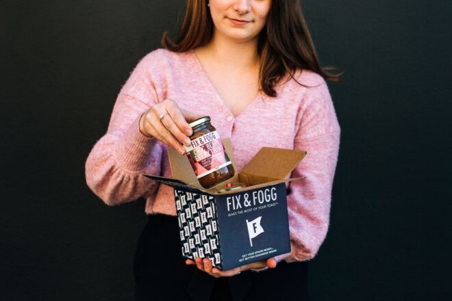 A person wearing a long-sleeved purple top holds a jar in one hand and a dark blue cardboard box in the other hand