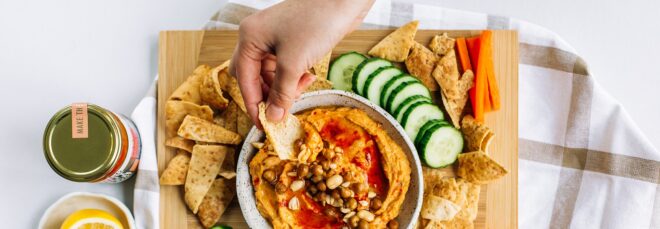 A white hand dipping a chip into a nut butter bowl filled with nuts. On the side of the bowl is more chips and slices of cucumbers